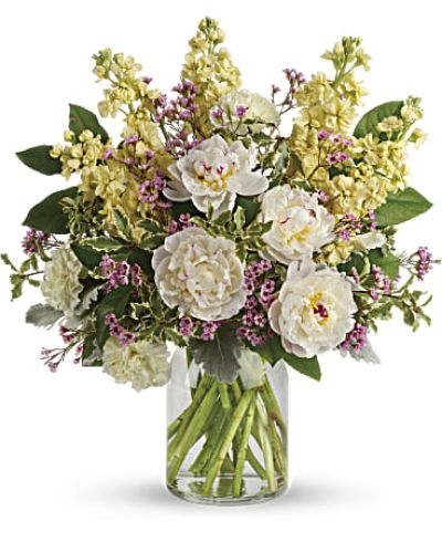 <p>At the heart of this bouquet are cloud-like white peonies, their lush petals opening with a sense of quiet grandeur. They are joined by pale yellow carnations, buttery-hued stock, and sprigs of lavender waxflower for a hint of color and texture. Silvery dusty miller and glossy lemon leaf frame the blooms, all arranged in a clear milk jug-style vase that balances rustic charm with refined elegance.</p>
