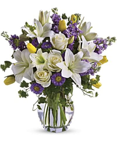 <p>A soft blend of white Asiatic lilies, creamy roses, golden tulips, and shades of lavender stock and matsumoto asters gives the bouquet its timeless charm. Accents of bupleurum, ivy, and seeded eucalyptus add natural depth, while a tall clear glass vase enhances the airy, flowing silhouette of the arrangement.</p>